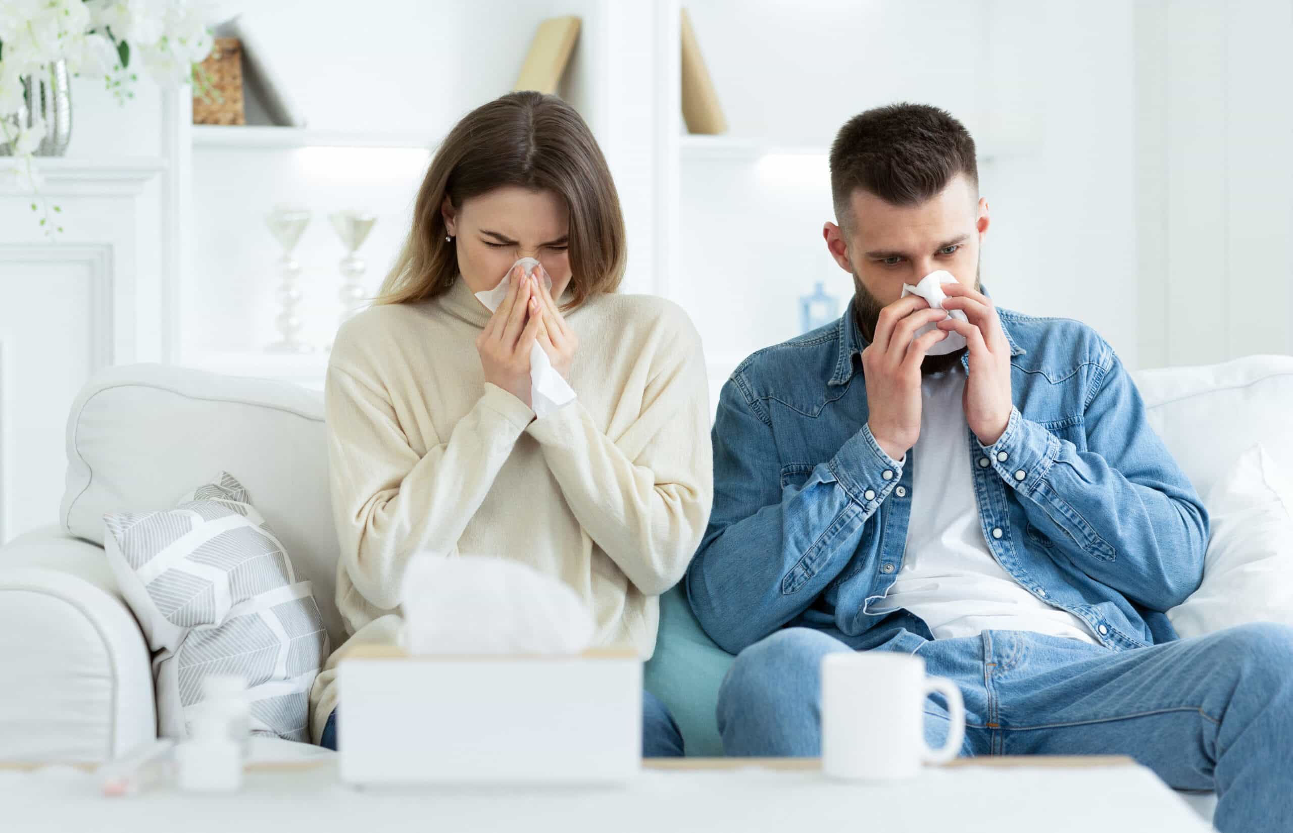 a man and woman sitting on a couch and blowing their noses due to allergies